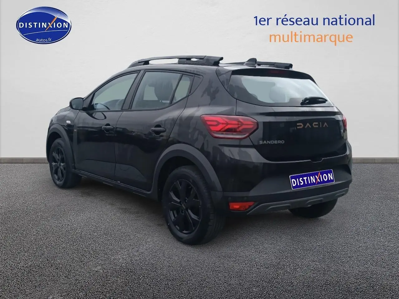 Vue 3/4 arrière droite d'une Dacia Sandero noire avec barres de toit et jantes alu noires, stationnée devant un mur clair.
