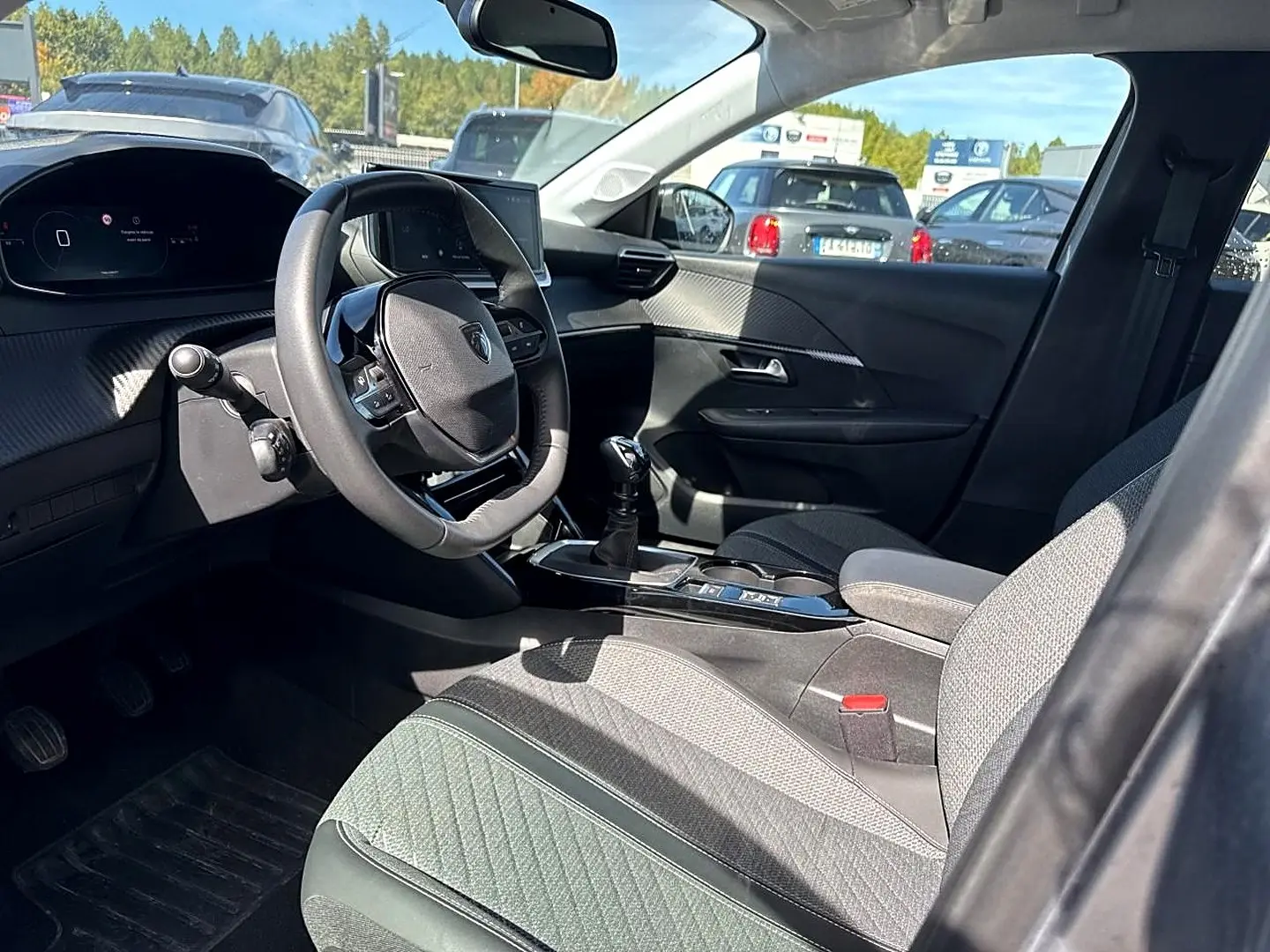 Vue intérieure côté conducteur de la Peugeot 208 1.2 100 Allure 2025, montrant le volant, la console centrale et les sièges tissu gris.