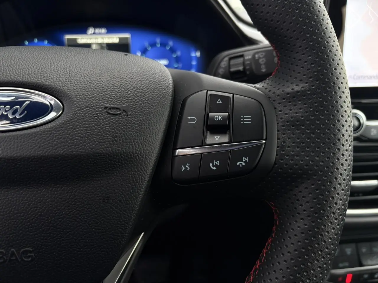 Gros plan sur le volant perforé noir avec surpiqûres rouges du Ford Puma 2024, boutons multifonctions visibles.