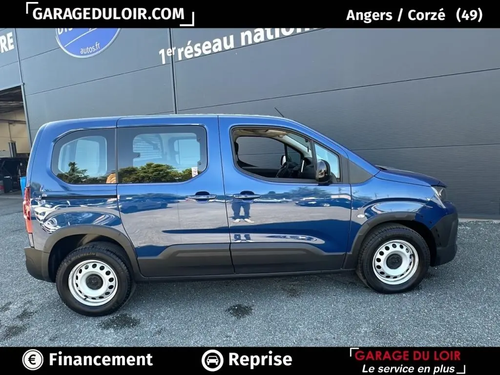 Vue latérale droite d’un Peugeot Rifter bleu 2024 avec roues acier et vitres teintées, stationné devant un mur gris.