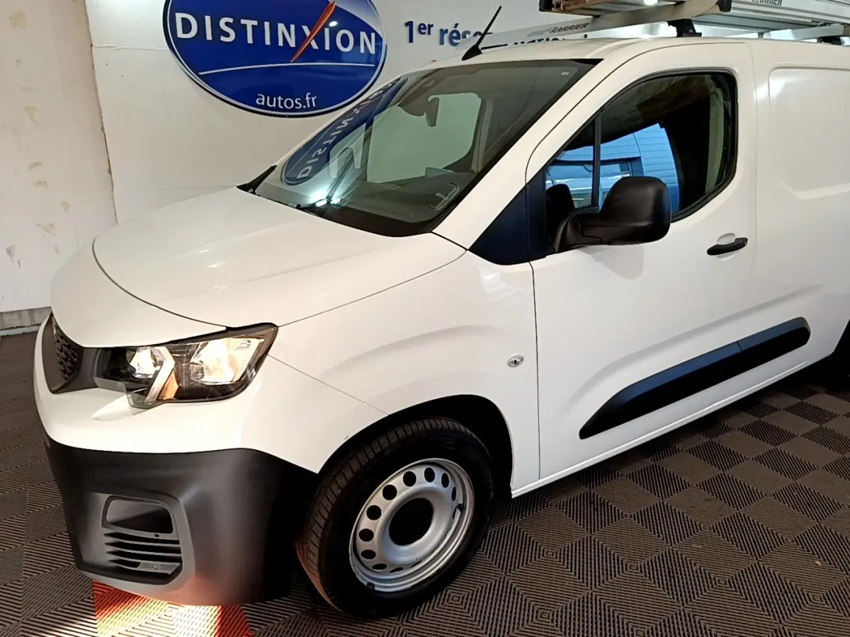 Vue 3/4 avant droit d’un Peugeot Partner blanc avec pare-chocs noir et barres de toit en intérieur showroom.