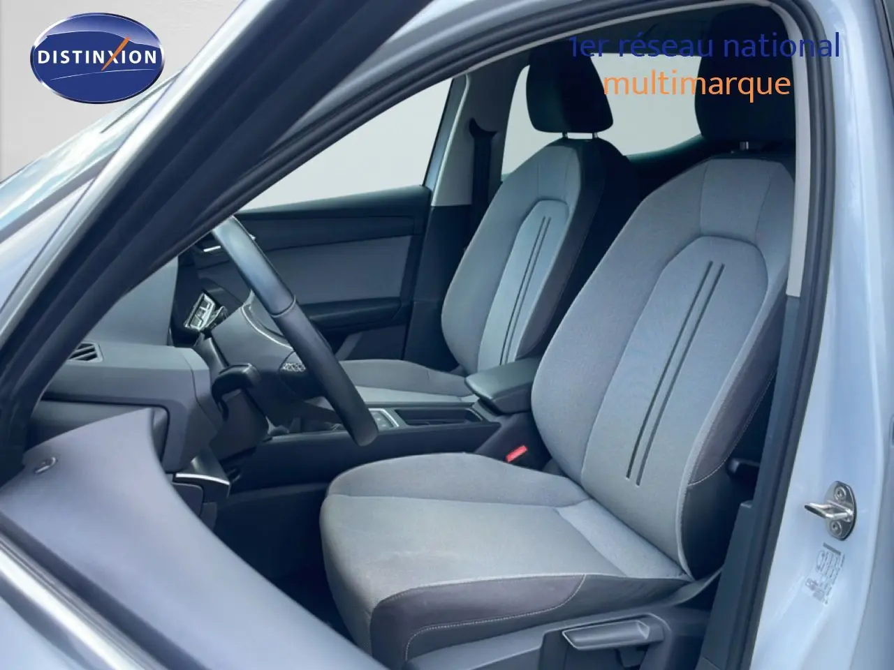 Vue intérieure côté conducteur d'une SEAT LEON 2025 blanc neige métal, sièges tissu gris clair avec détails noirs.