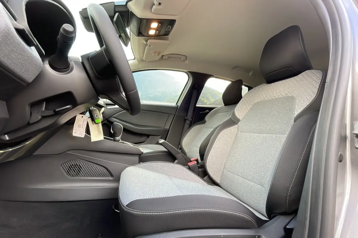Vue intérieure côté conducteur de la Renault Clio 2025, sièges tissu gris clair avec surpiqûres noires et volant multifonctions.