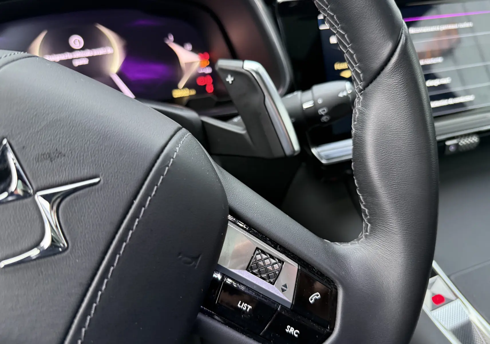 Gros plan sur le volant en cuir noir du DS7 2025, avec commandes intégrées et tableau de bord numérique flou en arrière-plan.