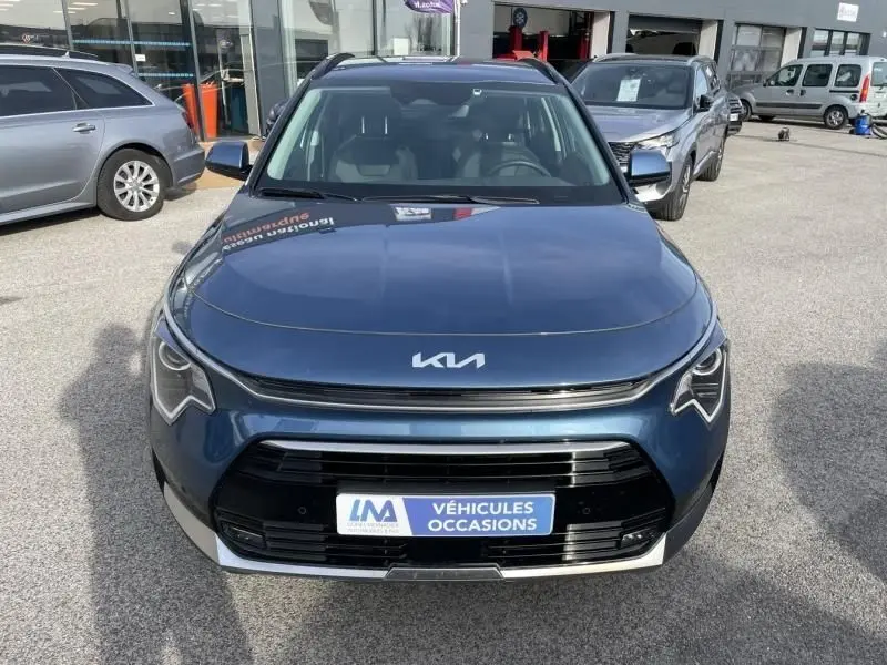Vue frontale d’un KIA Niro bleu métal 2024 avec calandre noire et logo KIA moderne, en extérieur sur parking.