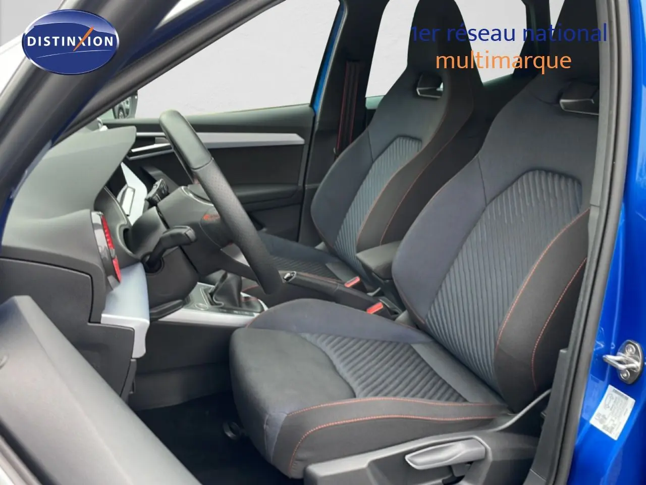 Vue intérieure côté gauche du SEAT Arona bleu saphir, montrant les sièges sport noirs avec surpiqûres orange et volant multifonction.