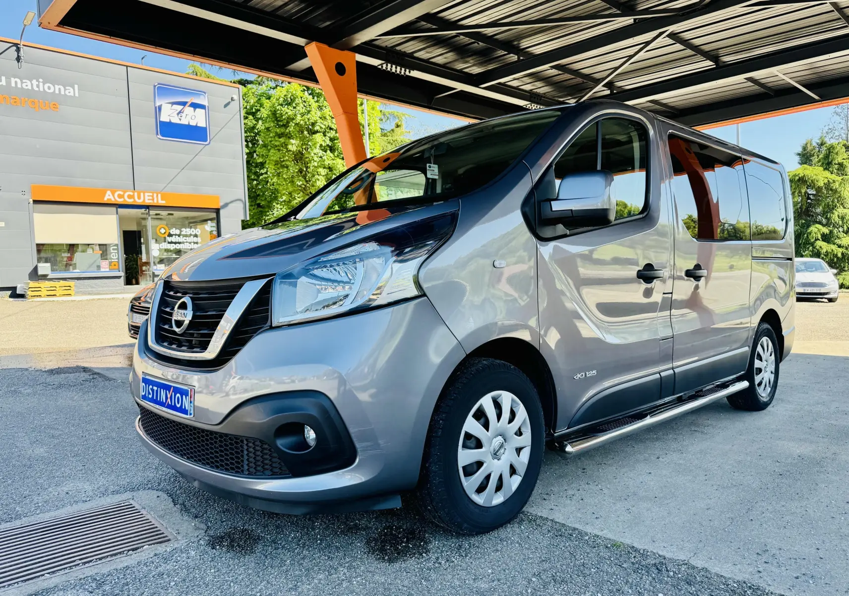 Vue 3/4 avant droit d’un Nissan NV300 Combi gris métallisé garé sous un auvent avec calandre noire et jantes tôles.