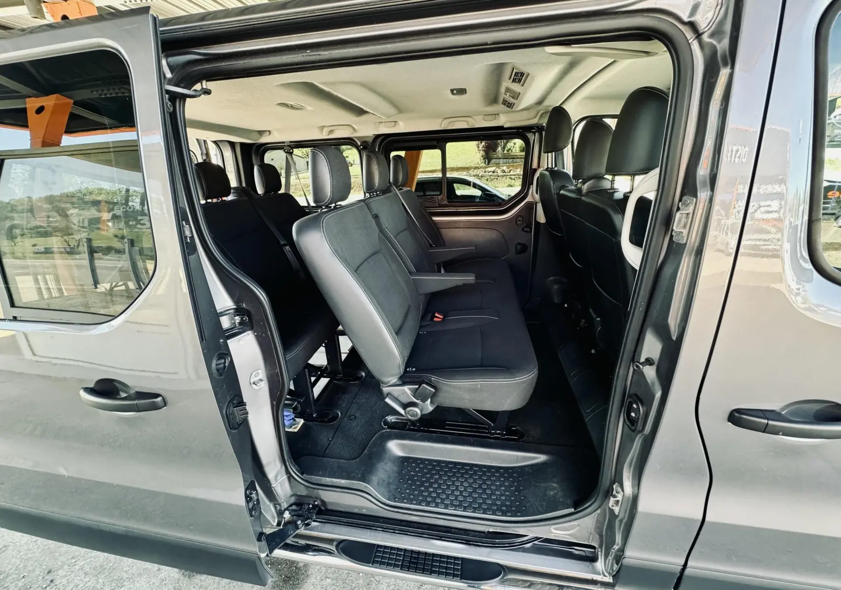 Vue latérale intérieure du Nissan NV300 Combi gris, montrant les sièges en tissu noir et la porte coulissante ouverte.