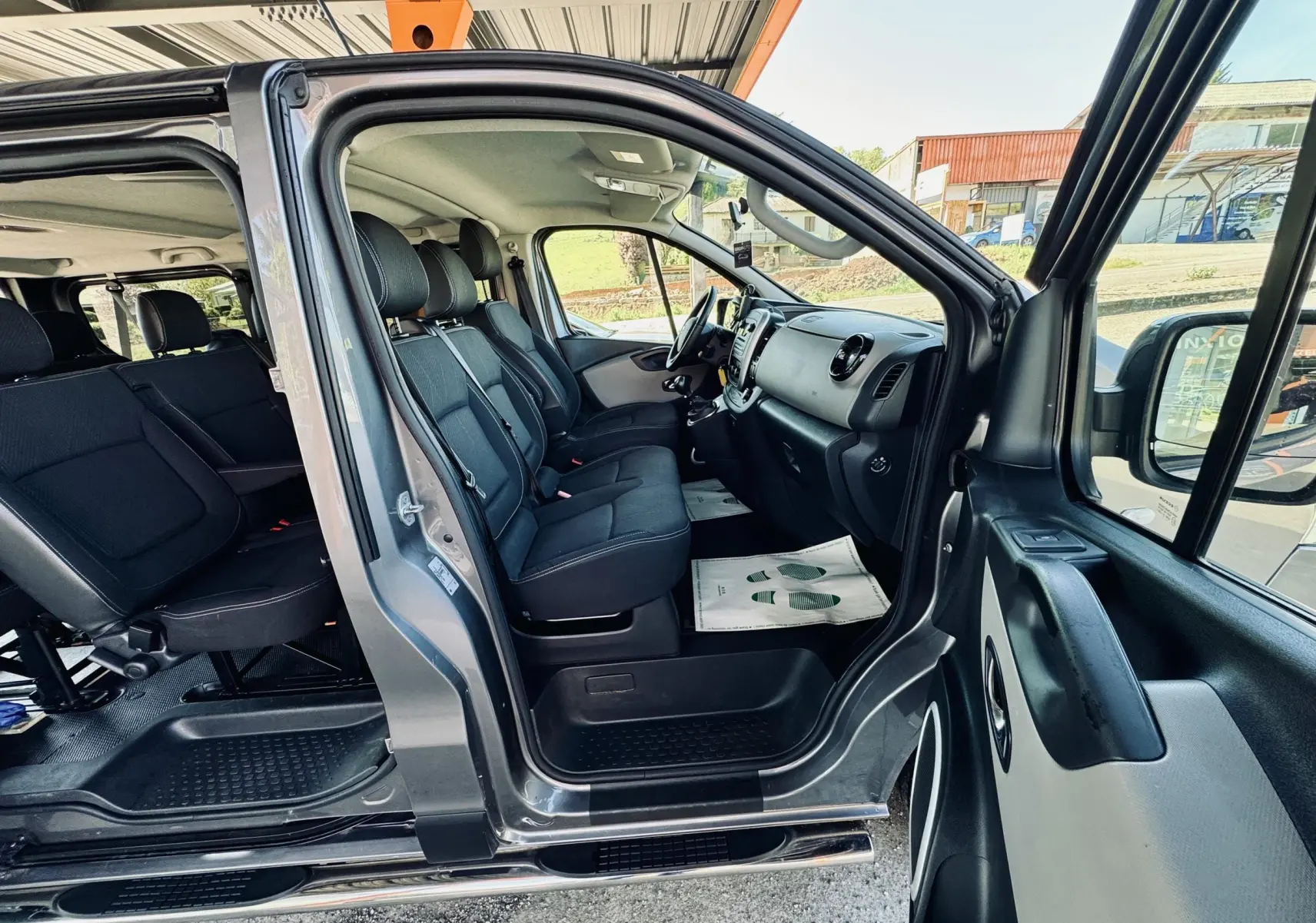 Vue côté gauche intérieur du Nissan NV300 Combi gris, montrant les sièges avant et arrière en tissu noir.