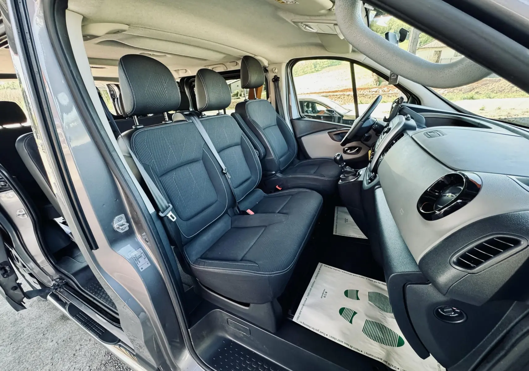 Vue intérieure côté conducteur du Nissan NV300 Combi gris, montrant les sièges en tissu noir et le tableau de bord.