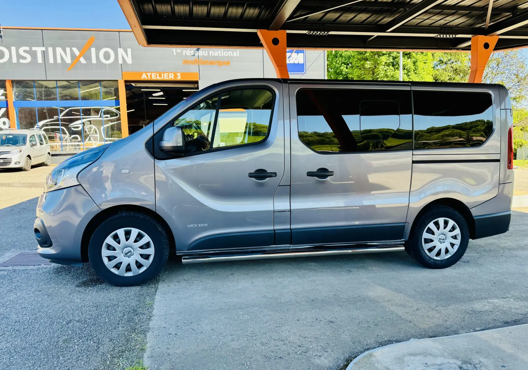 Profil latéral droit d’un Nissan NV300 Combi gris, 9 places, avec vitres arrière teintées, sous un abri couvert.