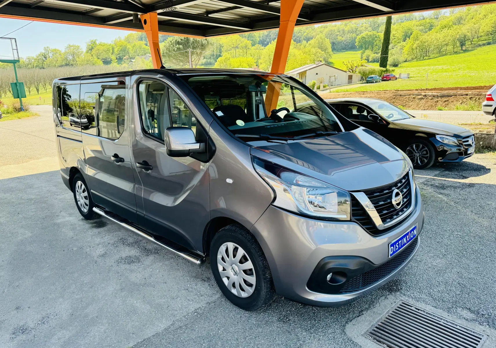 Nissan NV300 Combi gris en vue 3/4 avant droit sous abri avec paysage verdoyant en arrière-plan.