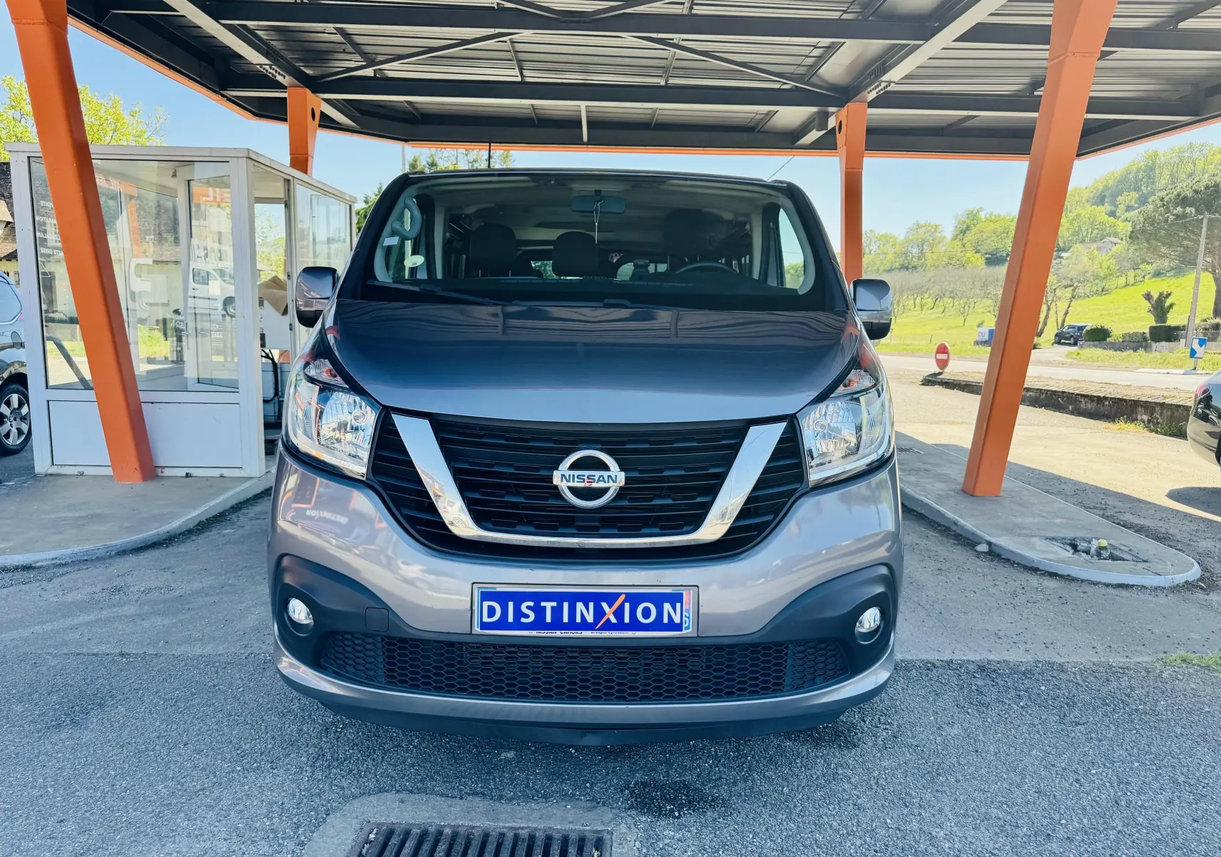 Vue frontale d’un Nissan NV300 Combi gris garé sous un abri avec plaque Distinxion visible.