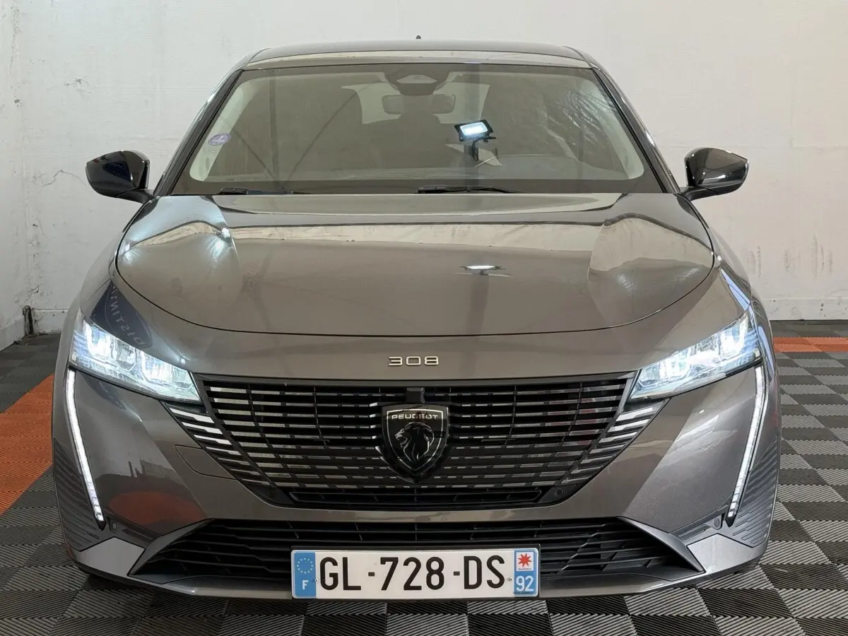 Vue frontale d’une Peugeot 308 gris foncé 2022 avec phares LED allumés et calandre noire distinctive.