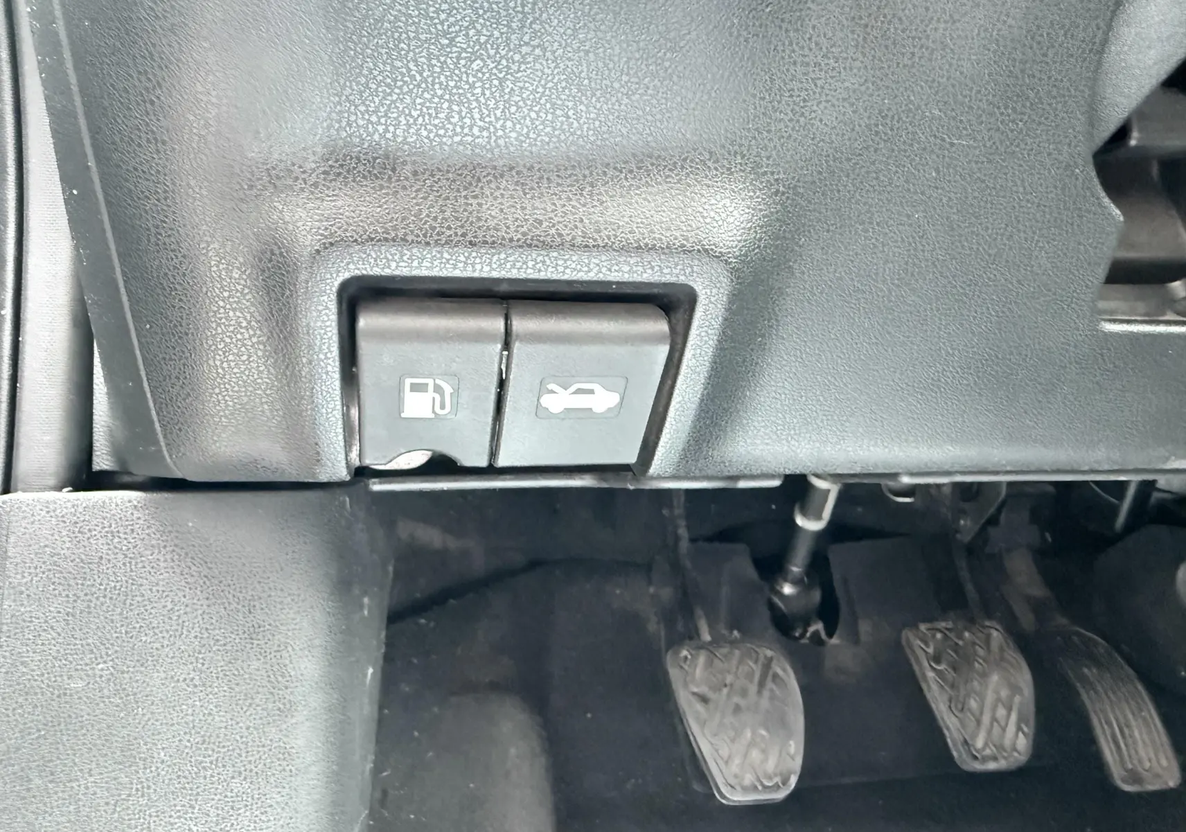 Gros plan sur les commandes d'ouverture de trappe à carburant et capot, avec pédales d'embrayage, frein et accélérateur visibles.