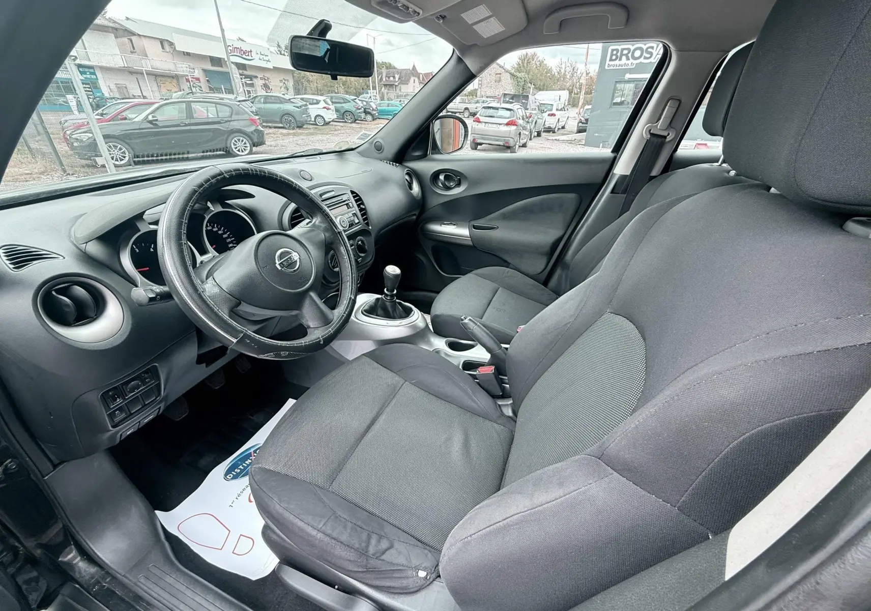 Vue intérieure côté conducteur du Nissan Juke noir 2012, montrant le volant cuir, la console centrale et les sièges tissu noirs.