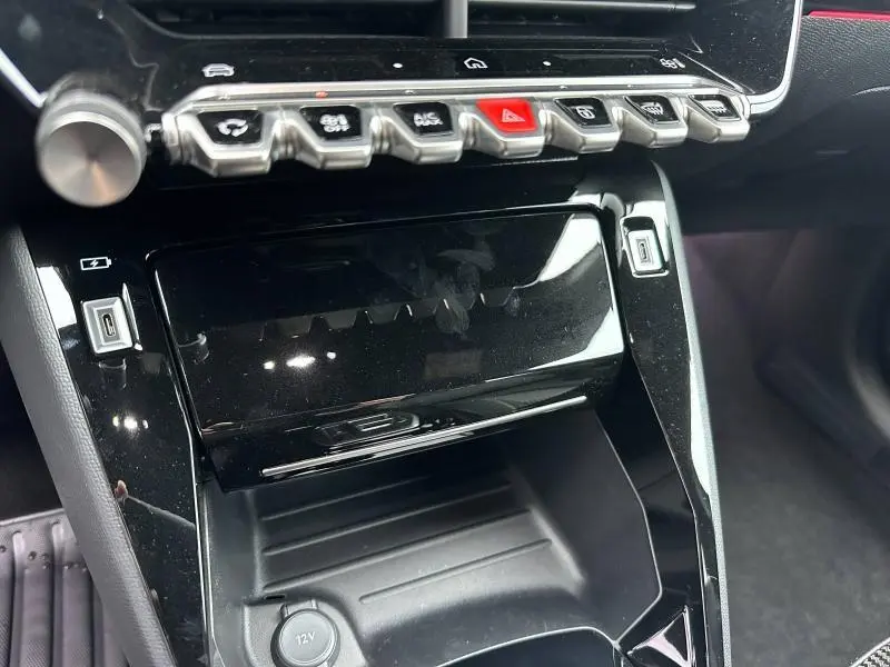 Gros plan sur la console centrale noire brillante de la Peugeot 208 Hybrid 2025 avec boutons de commande et prise USB visible.