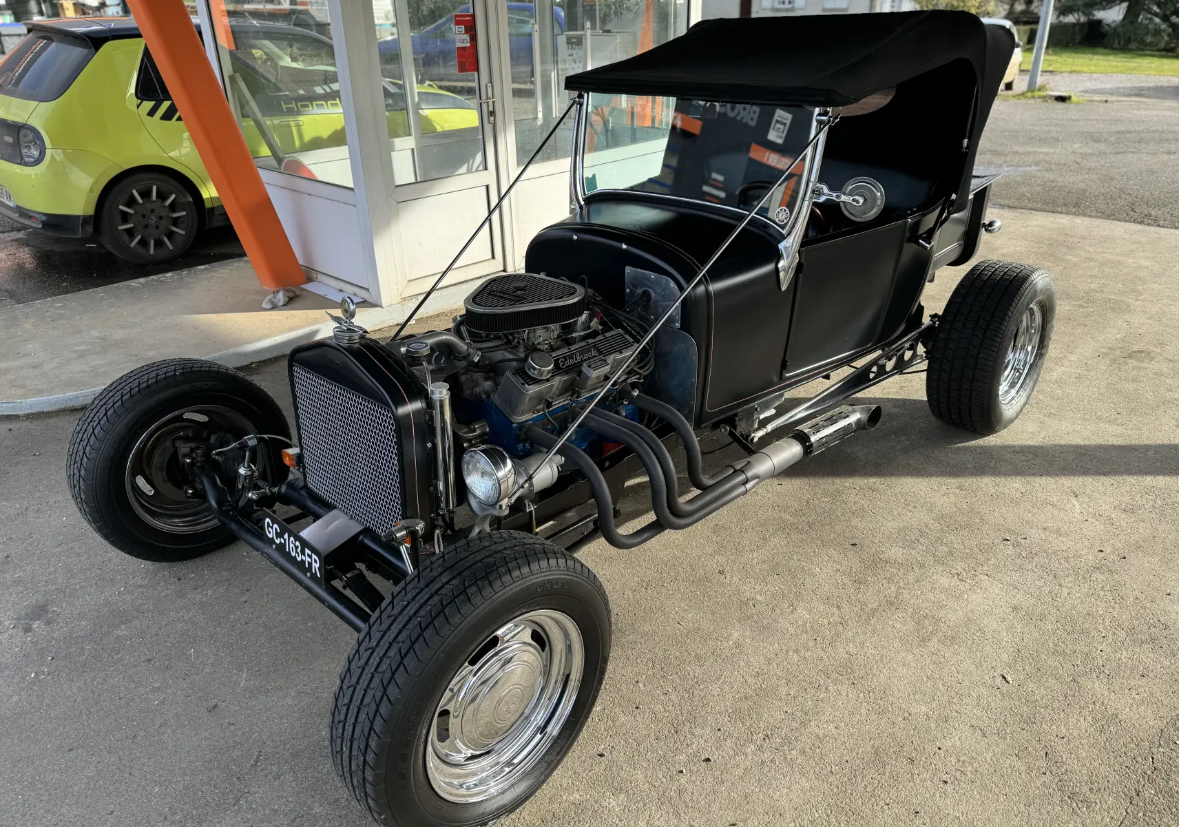 Ford T Hot Rod V8 noire vue 3/4 avant droit, capote relevée et moteur V8 apparent avec échappements latéraux.