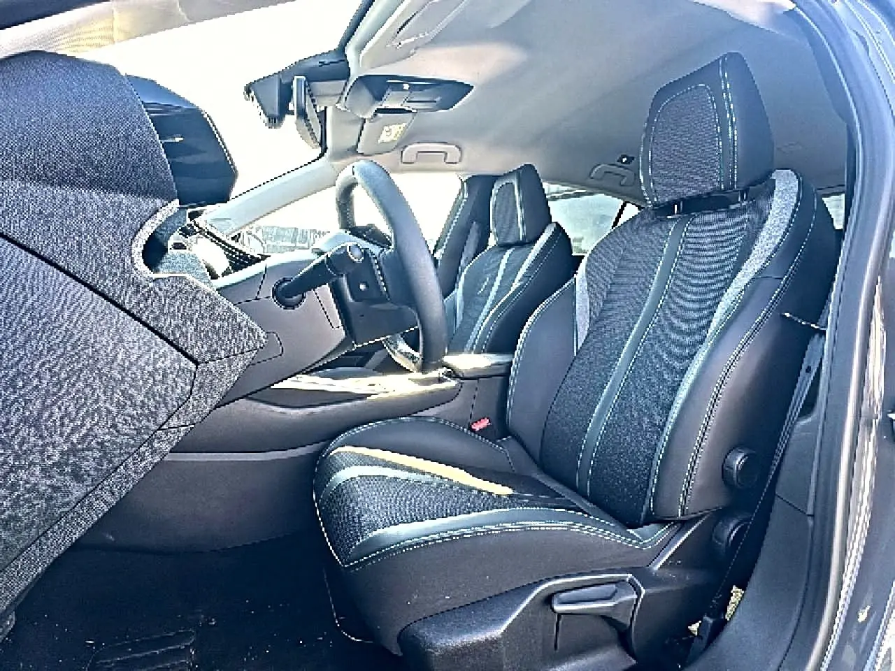 Vue intérieure côté conducteur de la Peugeot 308 2025, sièges tissu noir avec surpiqûres contrastantes et accoudoir central.
