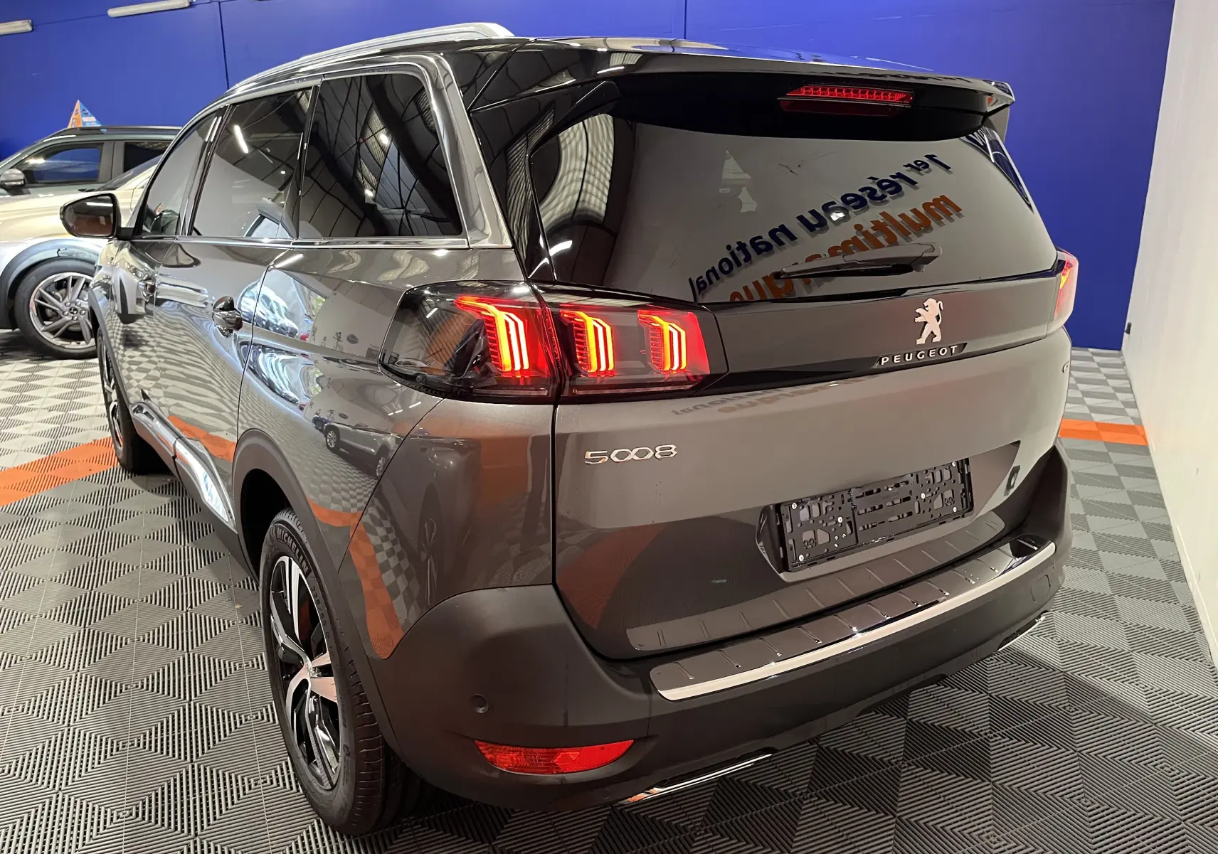 Vue 3/4 arrière droite du Peugeot 5008 gris platinium 2024 avec feux arrière LED allumés dans un showroom.