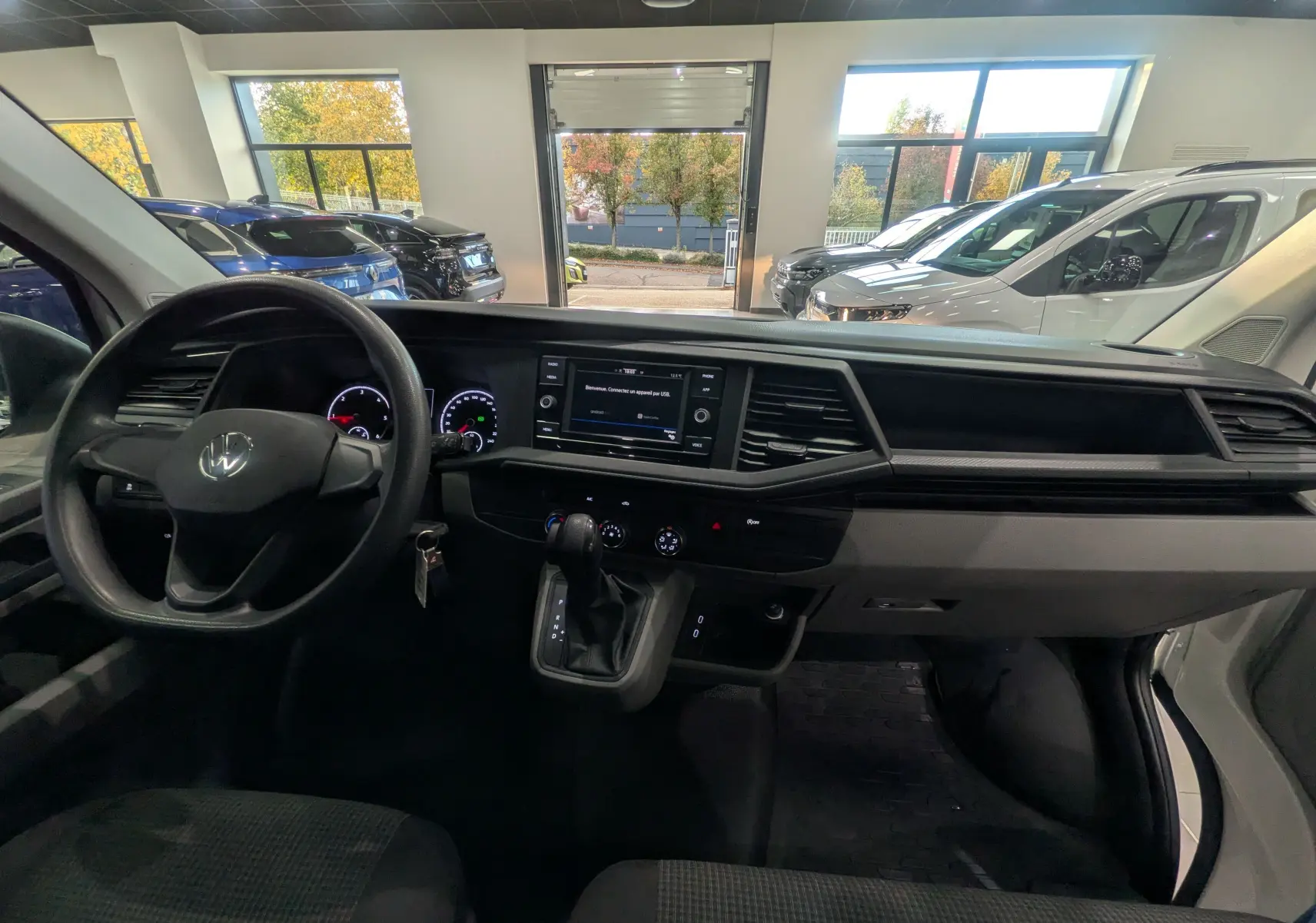 Vue intérieure du tableau de bord du Volkswagen Transporter 6.1 blanc, avec volant à gauche et écran central tactile allumé.