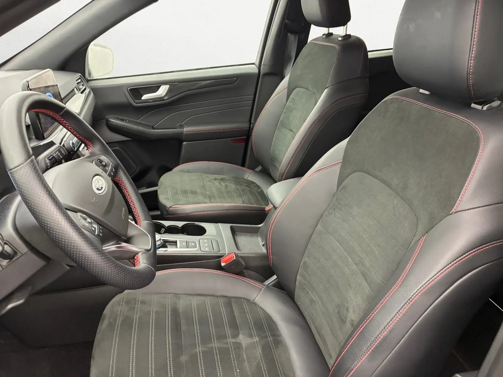 Vue intérieure côté conducteur du Ford Kuga 2024, sièges noirs en cuir et alcantara avec surpiqûres rouges, volant multifonction.