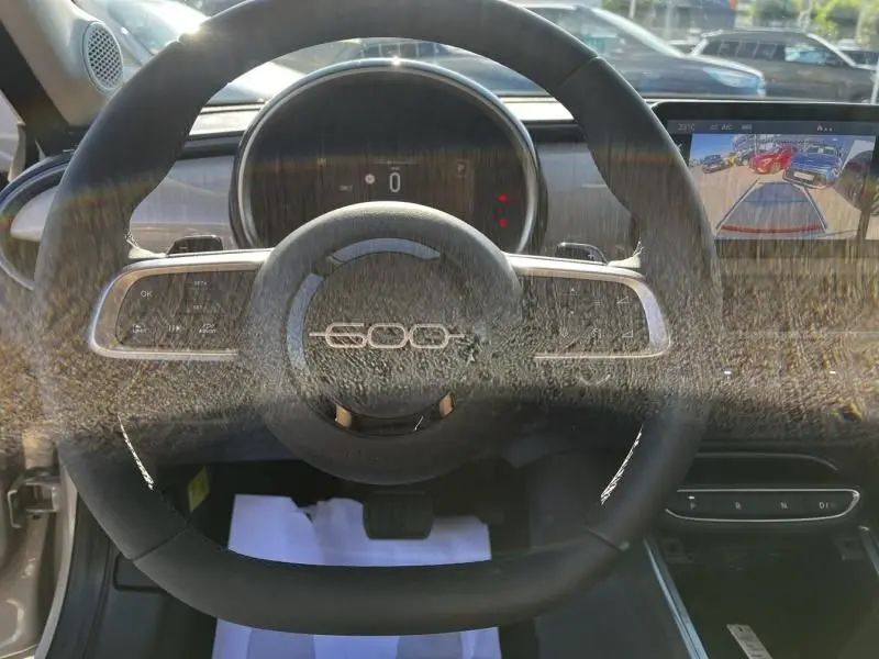 Vue intérieure centrée sur le volant noir avec logo 600, tableau de bord numérique et écran multimédia du Fiat 600 beige 2025.