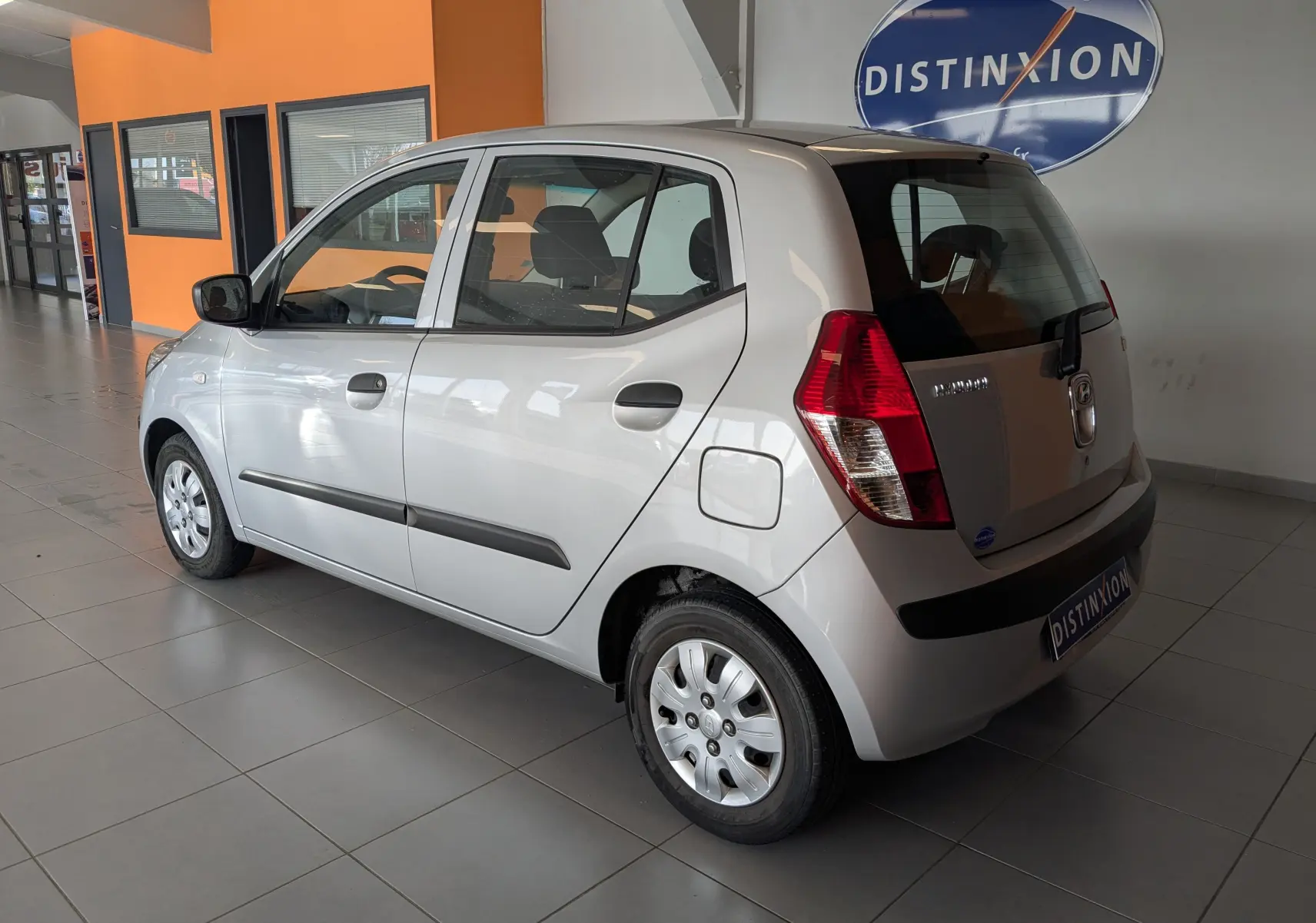 Vue 3/4 arrière droite d'une Hyundai i10 gris clair 5 portes, avec poignées noires et jantes acier en intérieur showroom.