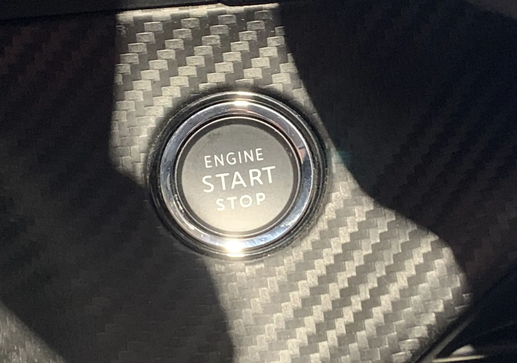 Gros plan sur le bouton start/stop moteur entouré d’un habillage effet carbone dans une Peugeot 208 gris, intérieur visible.