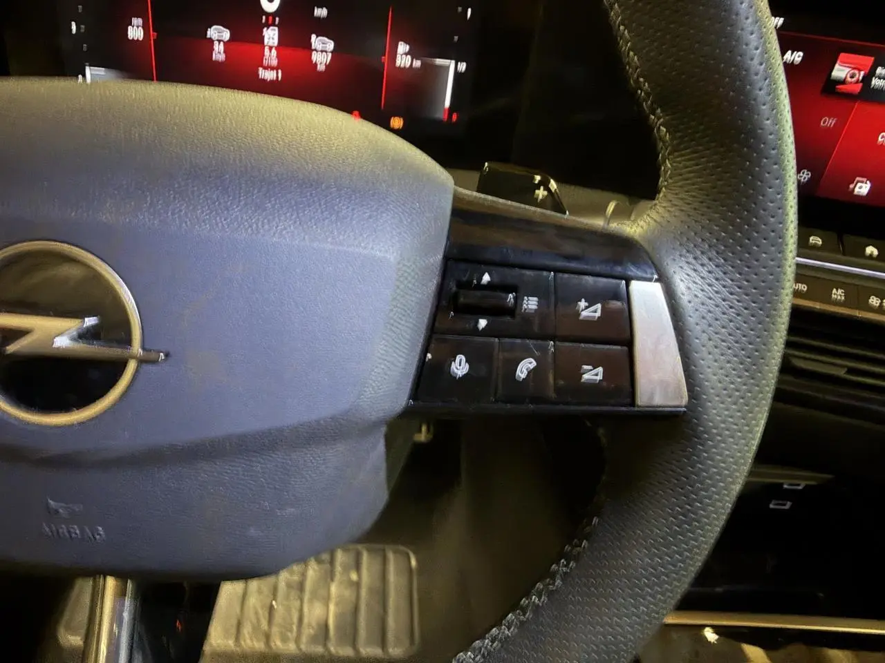 Gros plan sur le volant en cuir perforé de l’Opel Astra 2024, avec commandes multifonctions et tableau de bord digital rouge en arrière-plan.