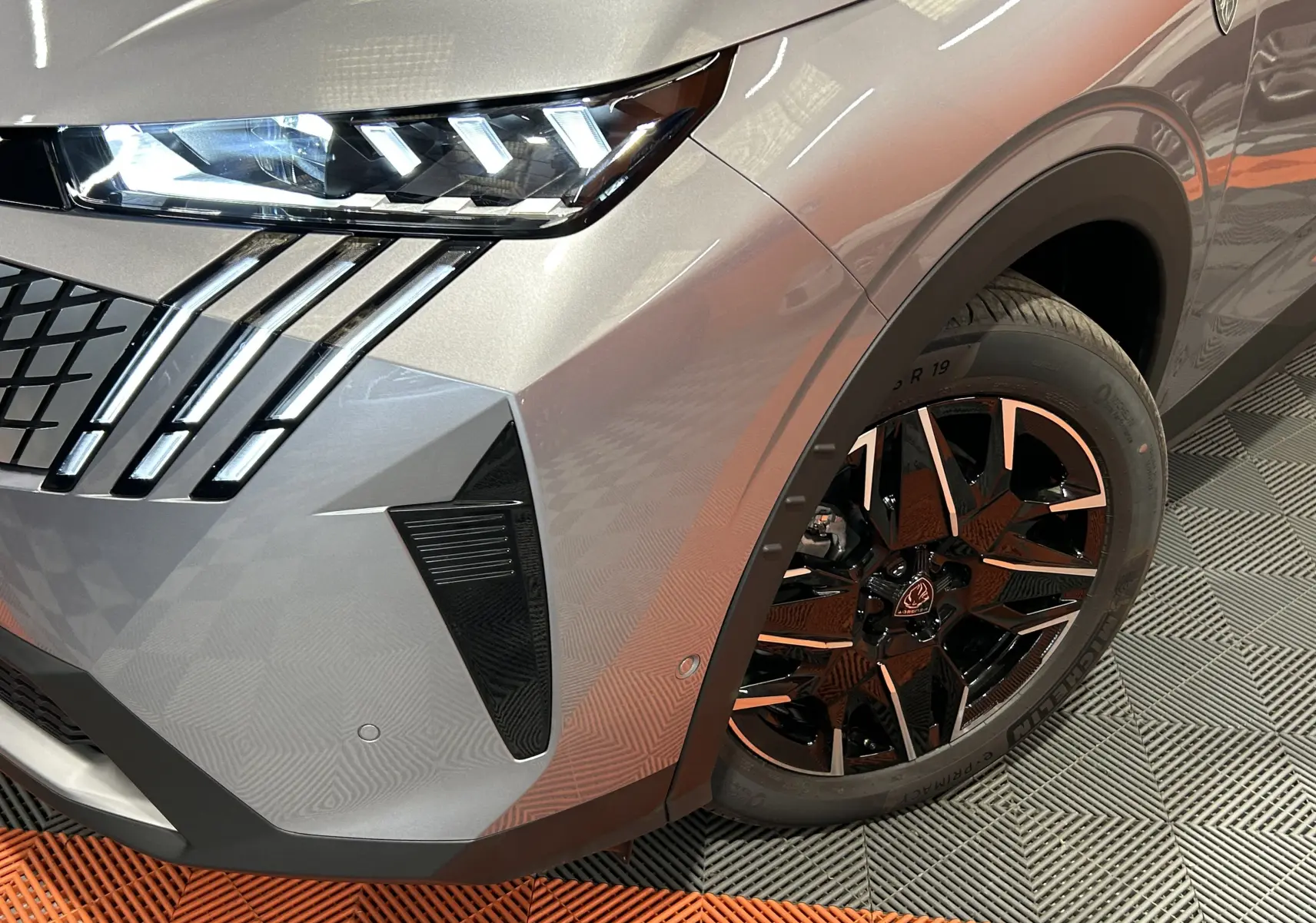 Gros plan sur l'avant droit gris Artense du Peugeot 5008 III GT Hybrid 145, mettant en valeur le phare LED et la jante biton.