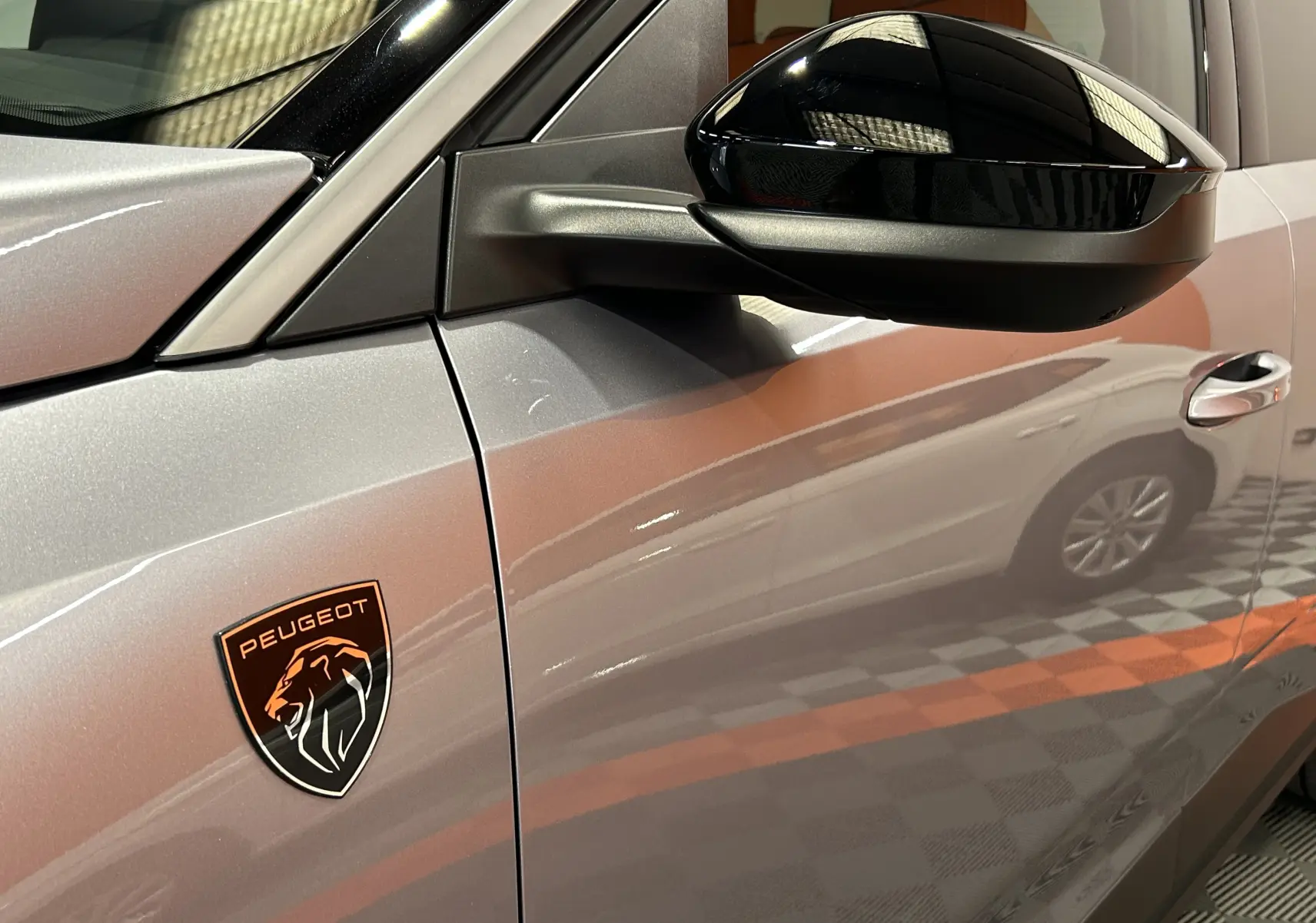 Gros plan sur le côté gauche gris Artense du Peugeot 5008 III GT Hybrid 145 avec logo lion noir et orange sur l'aile.