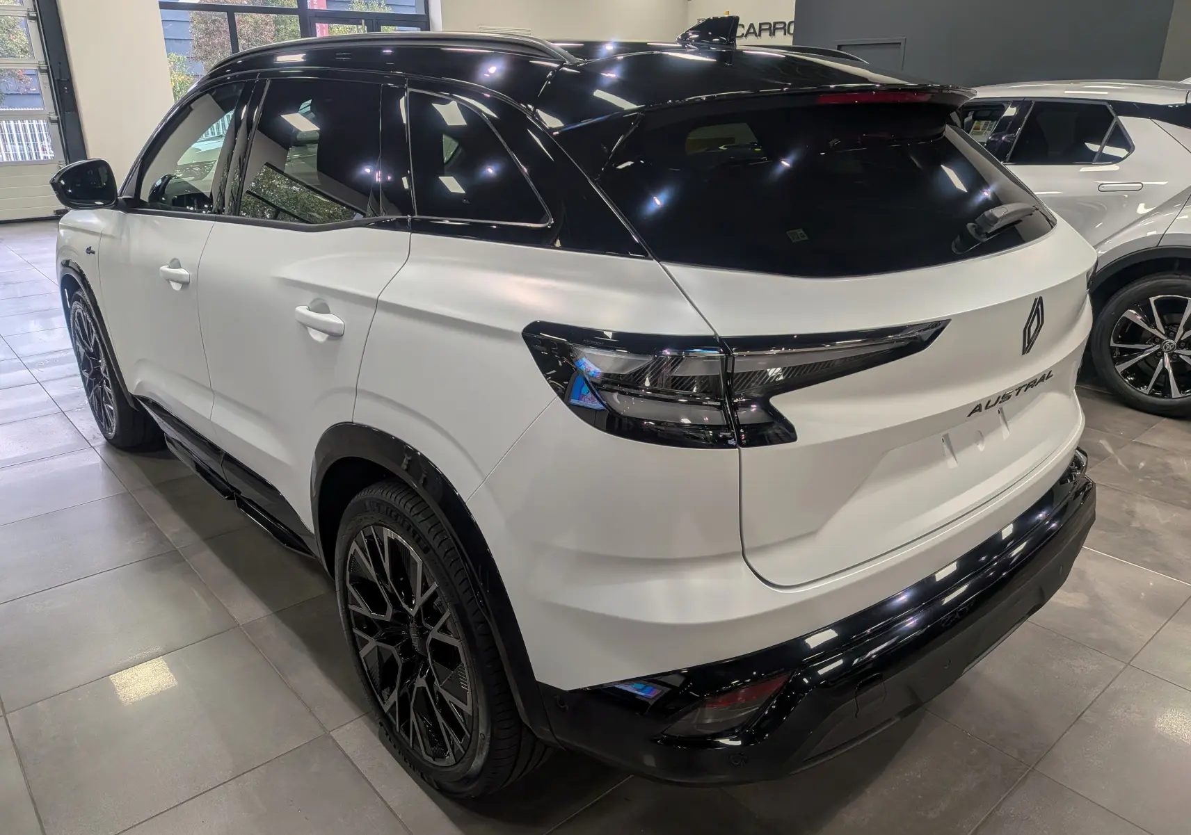 Vue 3/4 arrière droite du Renault Austral blanc nacré avec toit noir et jantes noires en intérieur showroom.