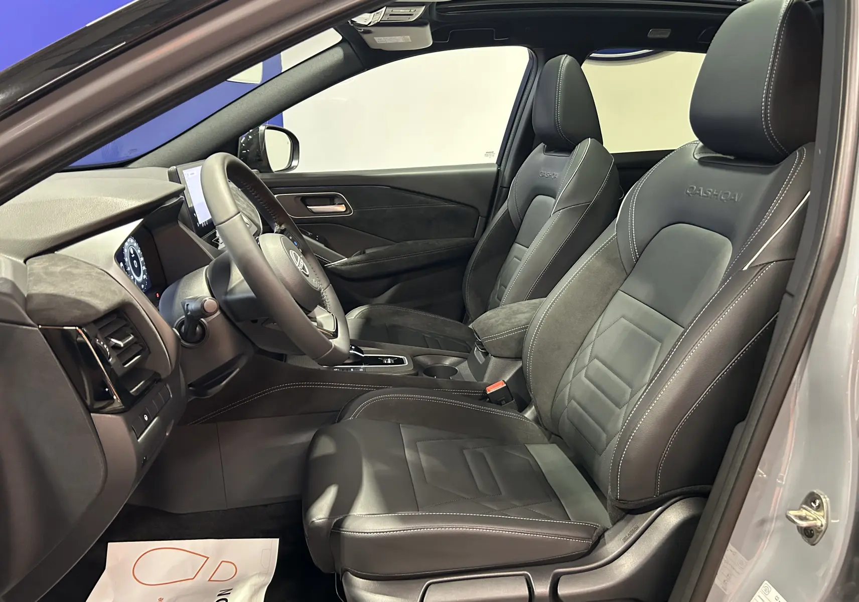 Vue intérieure côté conducteur du Nissan Qashqai gris céramique, sièges cuir noir avec logo Qashqai brodé et toit panoramique ouvert.