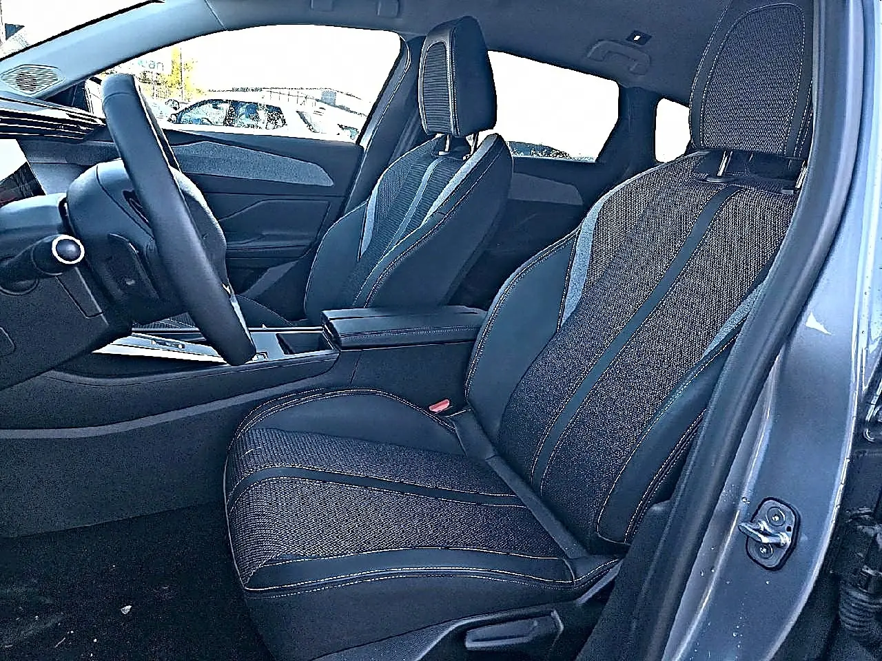 Vue intérieure côté conducteur de la Peugeot 308 SW 2025 gris Artense, avec sièges tissu et accoudoir central.