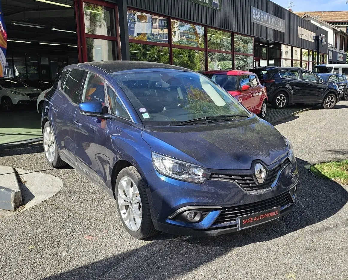 Renault Scenic bleu cosmos métal vu en 3/4 avant droit devant un garage AD Expert avec drapeau promotionnel.
