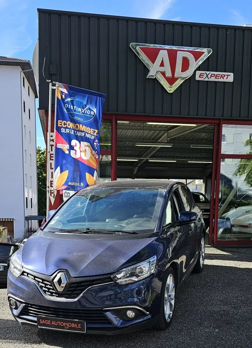 Renault Scénic bleu cosmos métal en 3/4 avant droit, garé devant un garage avec plaque SAGE AUTOMOBILE visible.