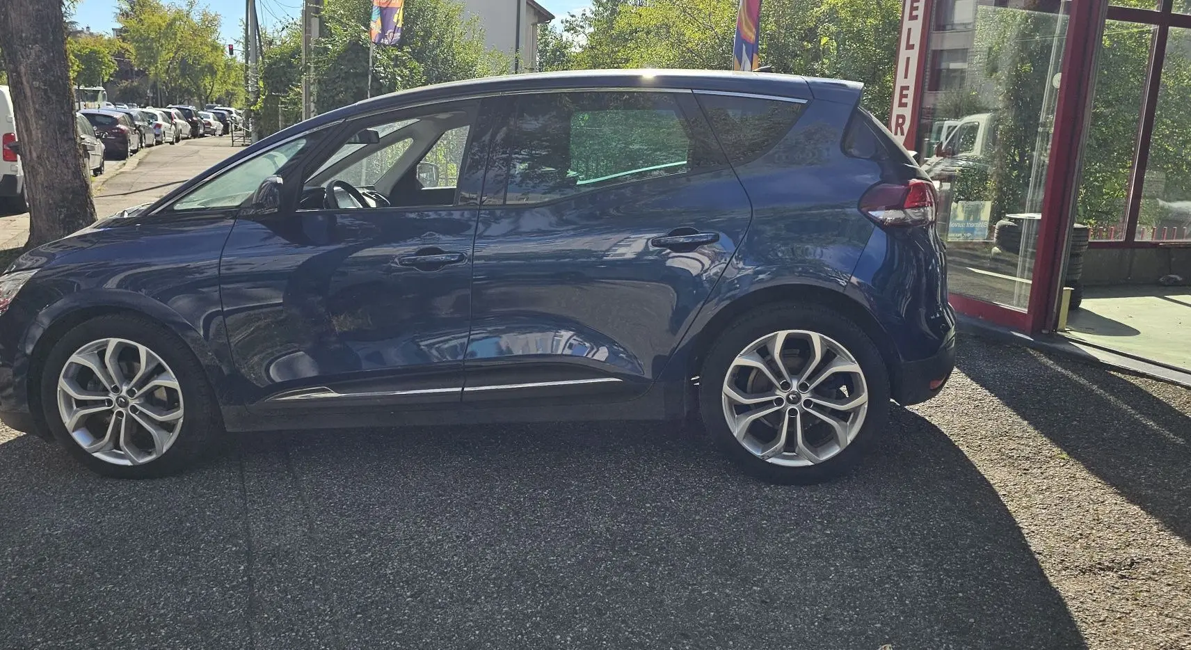 Profil côté gauche d'un Renault Scénic bleu cosmos métal 2020 garé devant un atelier avec jantes alliage visibles.