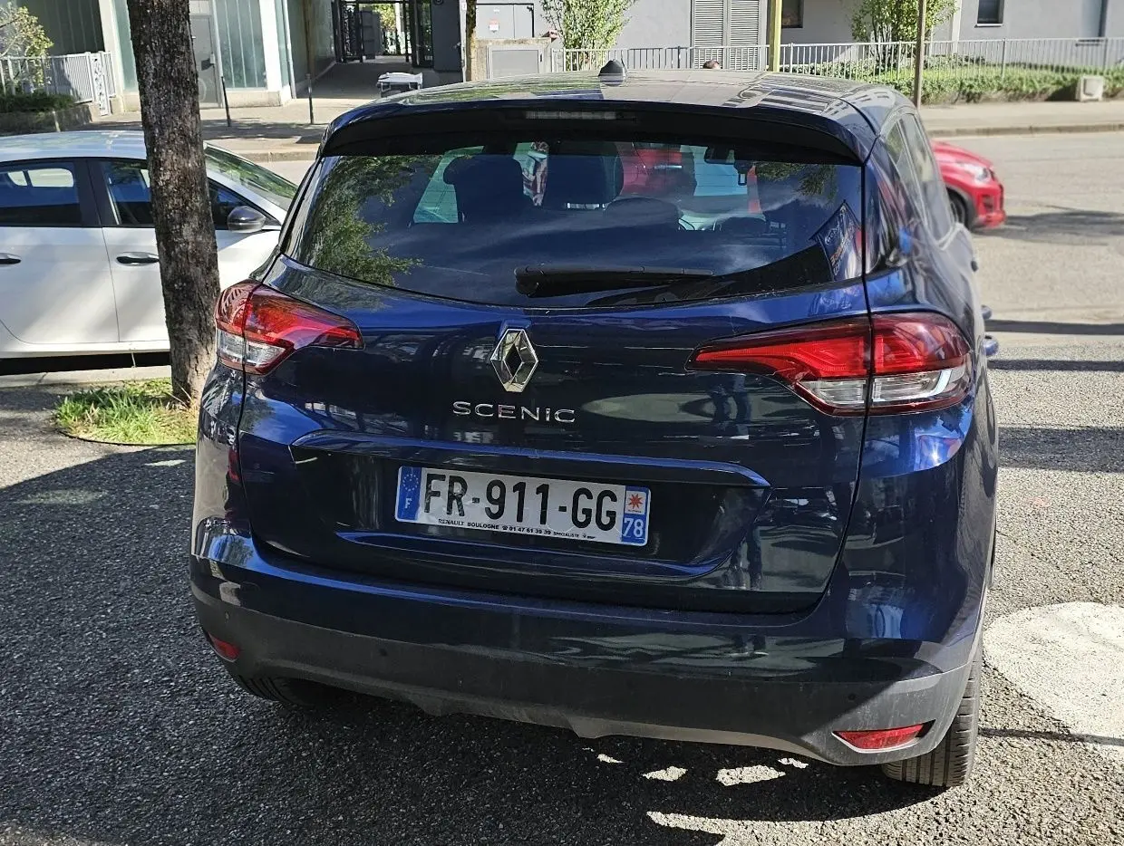 Vue arrière d'un Renault Scénic bleu cosmos métal garé, avec feux arrière rouges et plaque d'immatriculation française.