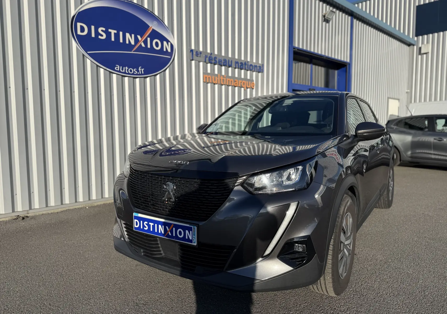 Peugeot 2008 gris 3/4 avant droit, avec calandre noire et phares LED, stationnée devant un bâtiment industriel.