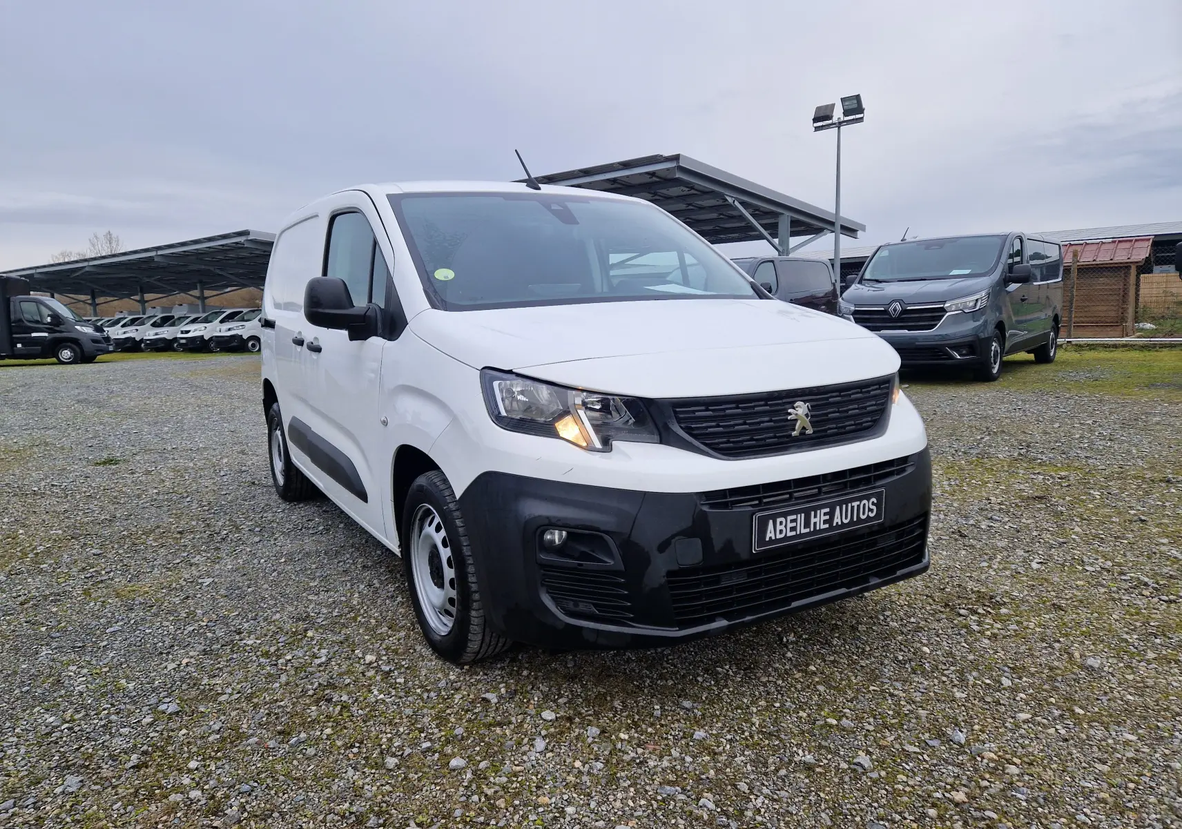 Peugeot Partner blanc vue 3/4 avant droit avec calandre noire et phare allumé sur parking extérieur.