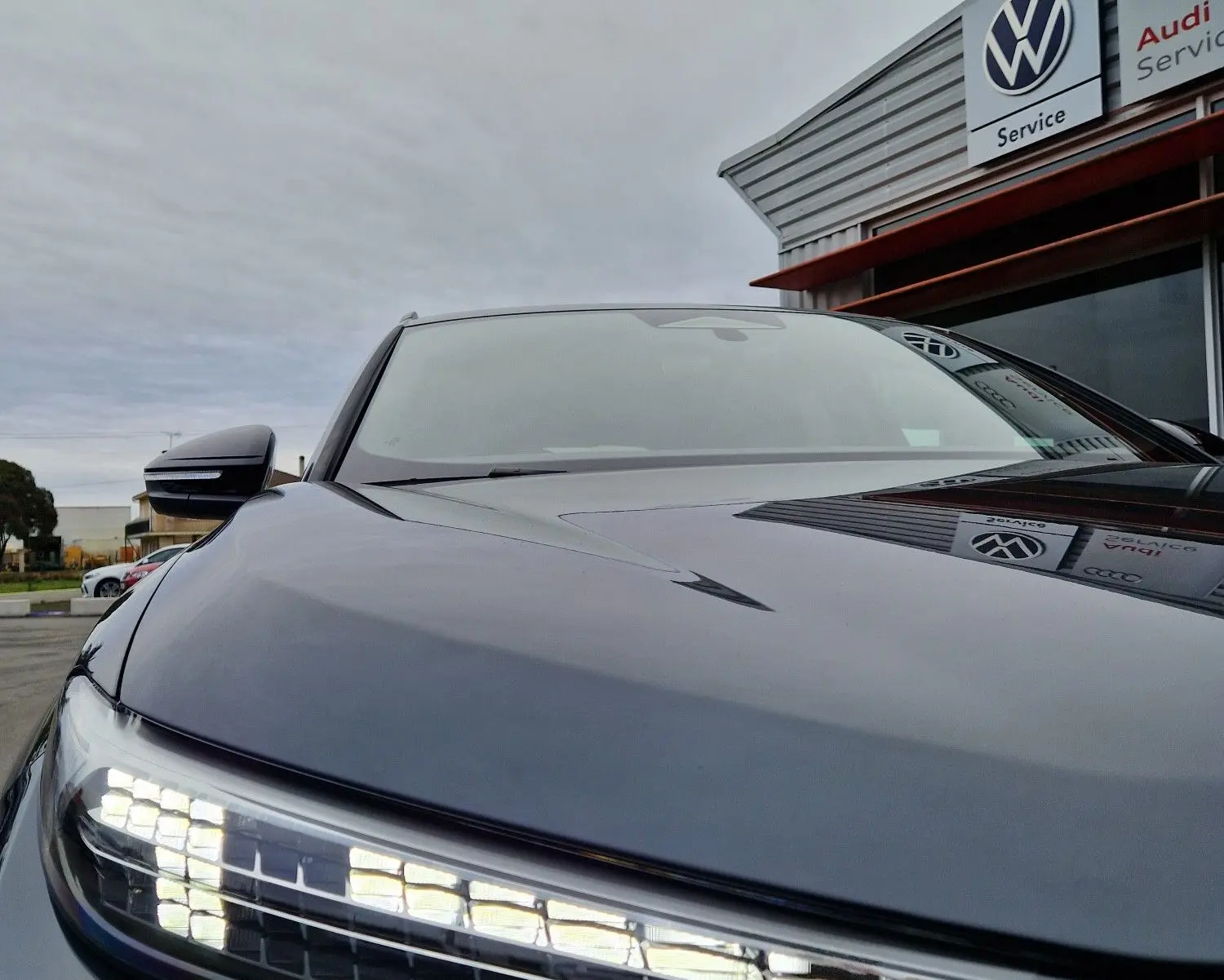 Gros plan sur le phare avant gauche allumé de l'Audi Q6 e-tron Performance S-Line gris Manhattan, avec reflet du ciel nuageux.