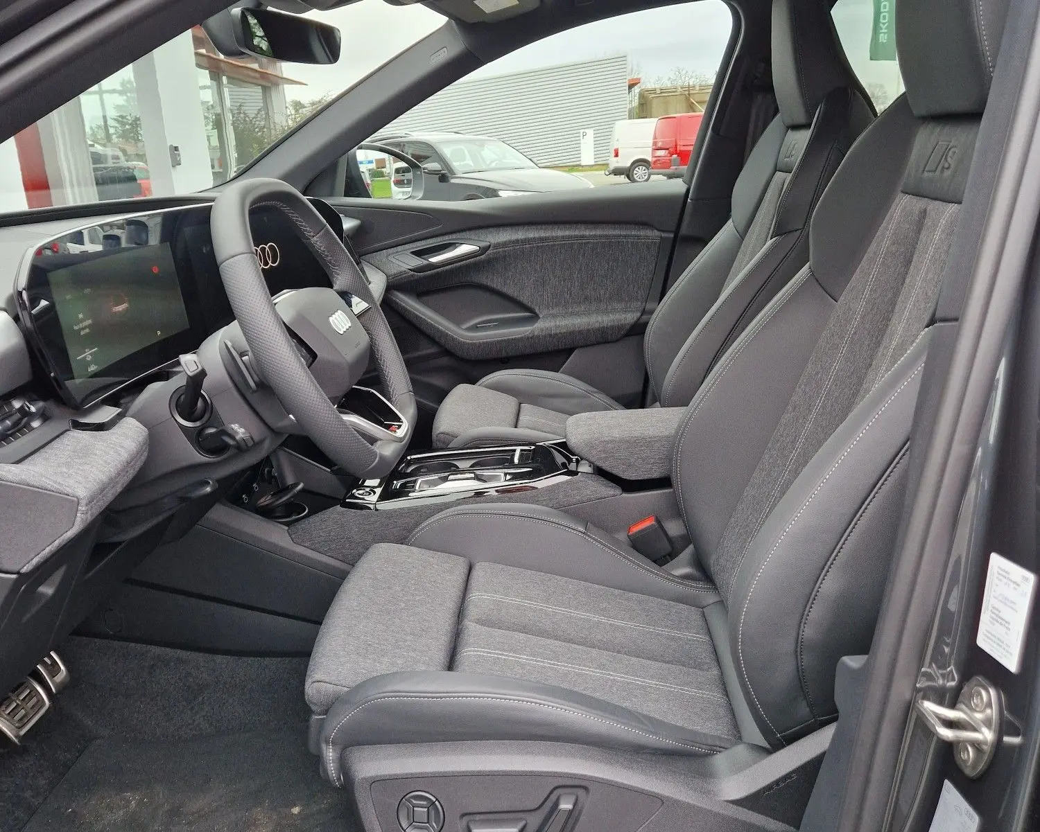 Vue intérieure côté conducteur de l'Audi Q6 e-tron Performance S-Line 2025 gris Manhattan, sièges sport avec logo S et tableau de bord digital.
