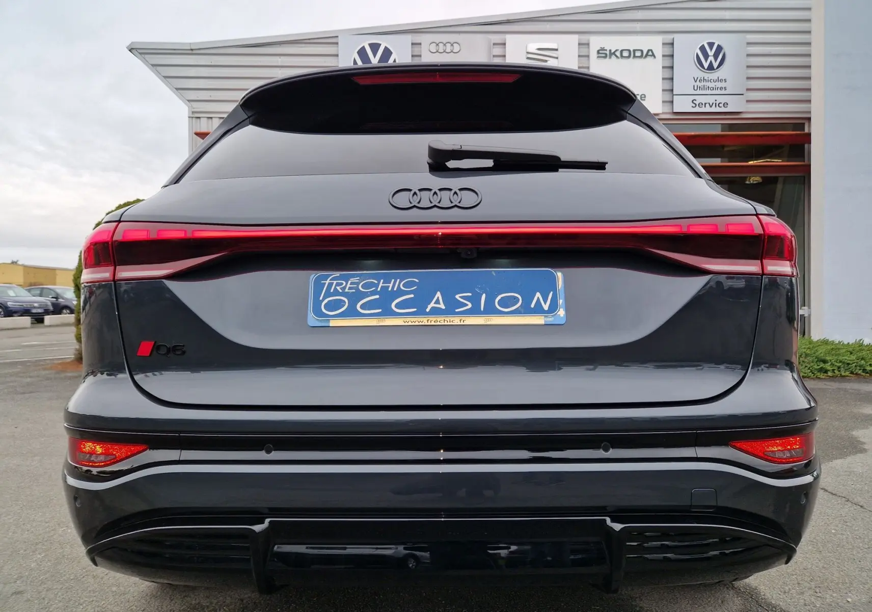 Vue arrière d’un Audi Q6 e-tron Performance S-Line gris Manhattan avec feux arrière LED continus et logo Q6 rouge.