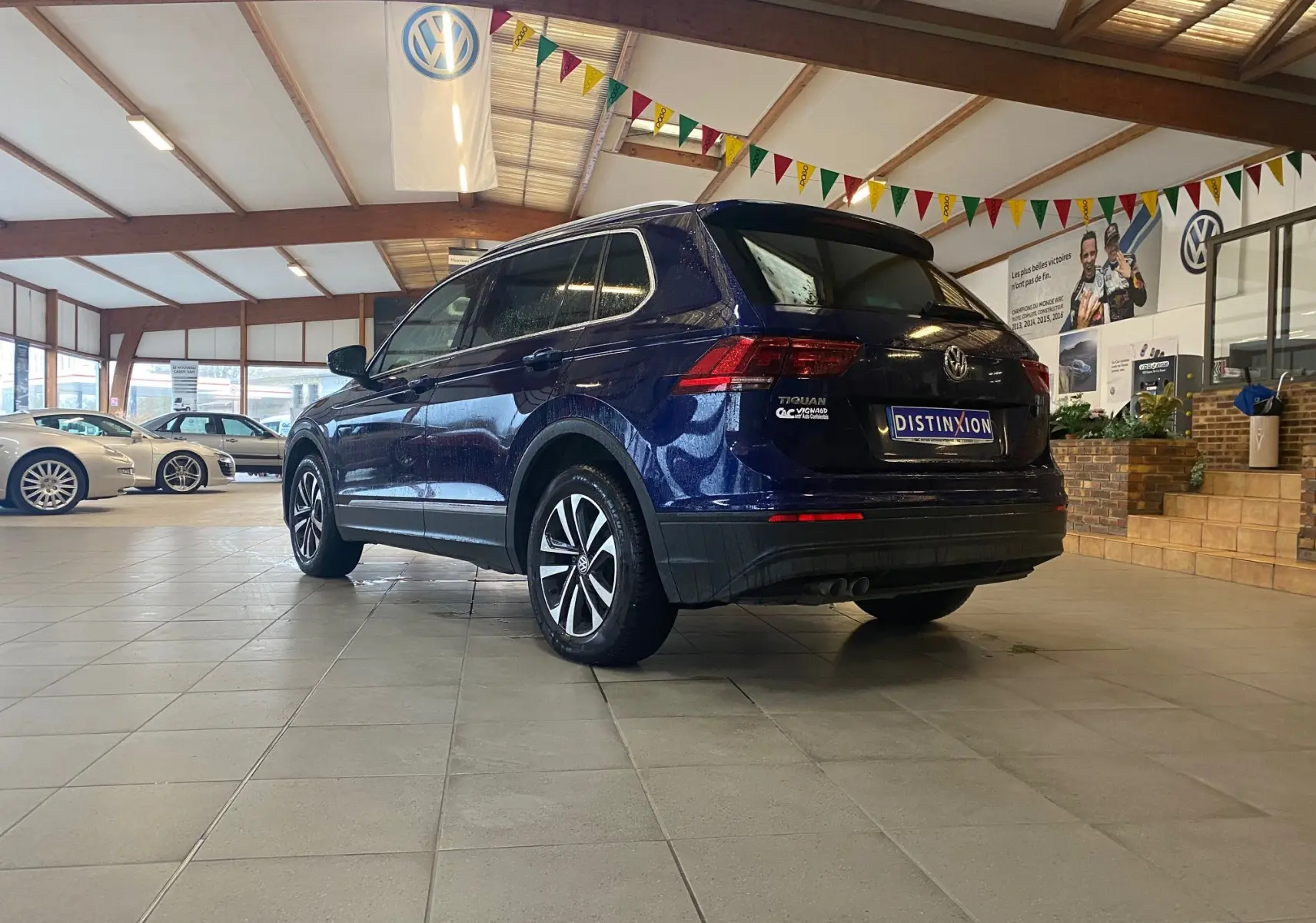 Volkswagen Tiguan bleu vert vu en 3/4 arrière droit dans un showroom avec drapeaux colorés suspendus.