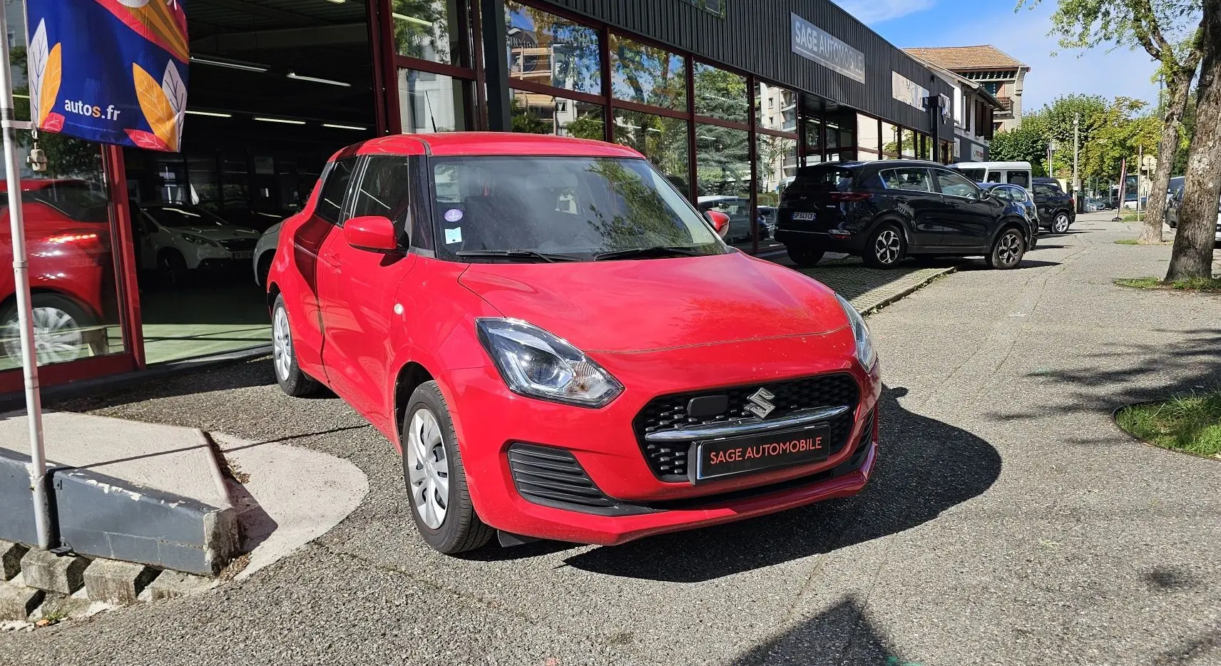 Suzuki Swift 2023 rouge fervent vue 3/4 avant garée devant un garage avec plaque Sage Automobile.