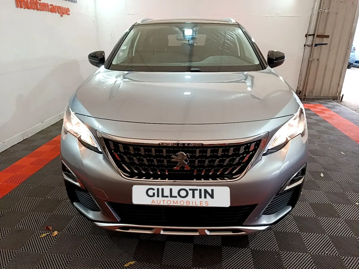 Vue avant d'un Peugeot 5008 gris clair 2020 avec calandre noire et phares allumés dans un garage.