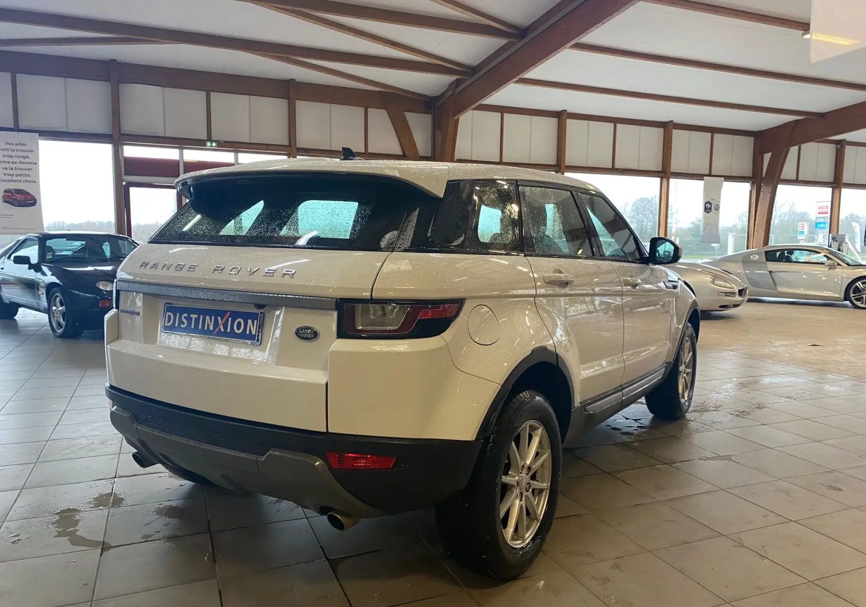 Vue 3/4 arrière droite d'un Range Rover Evoque blanc 2016 dans un showroom avec sol carrelé et autres véhicules visibles.