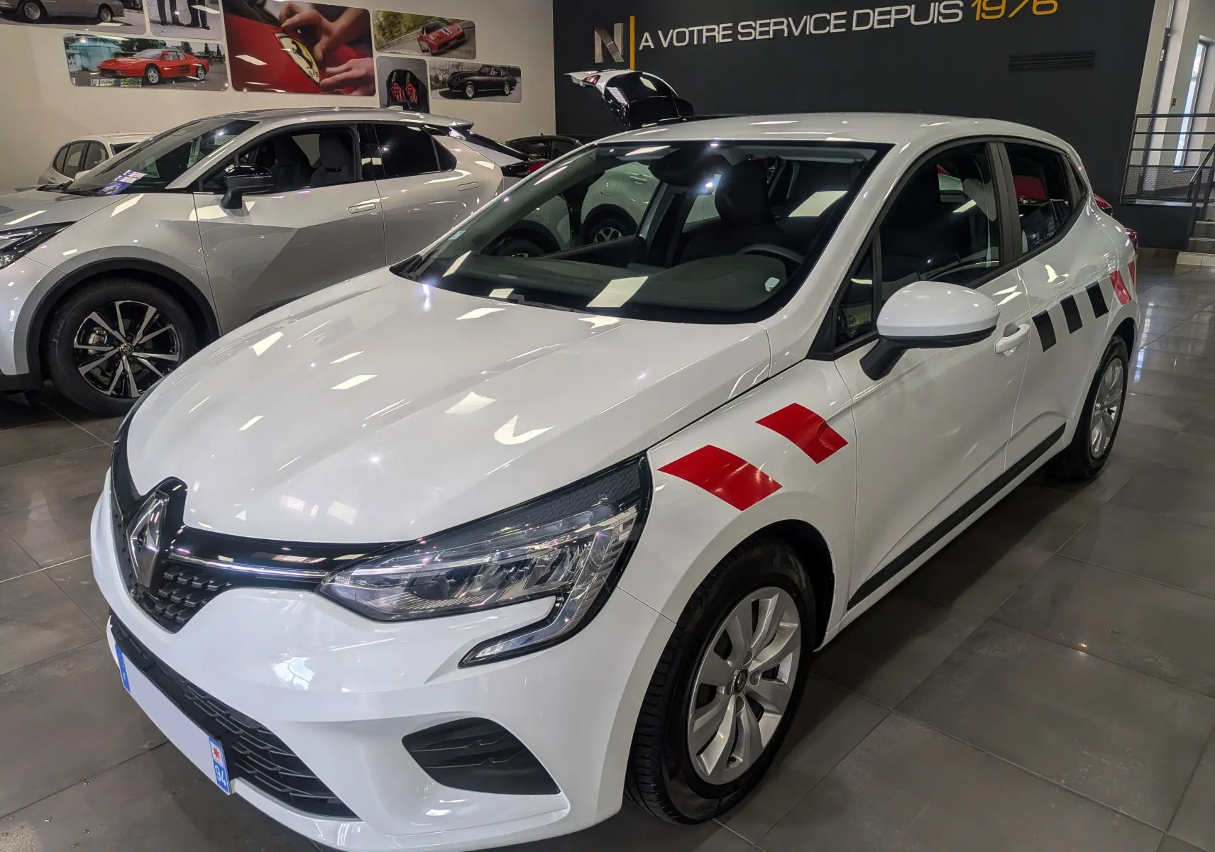 Renault Clio V blanche vue 3/4 avant droit avec marquages rouges sur ailes et porte dans un showroom.