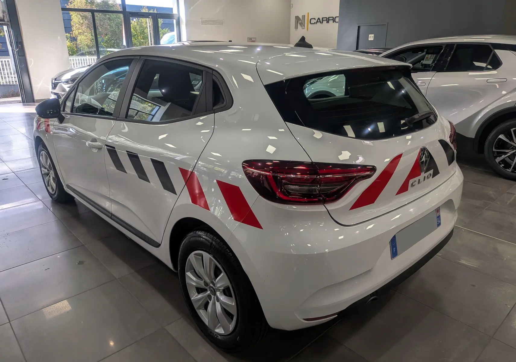 Vue 3/4 arrière droite d'une Renault Clio V blanche avec bandes rouges et noires distinctives sur la carrosserie.